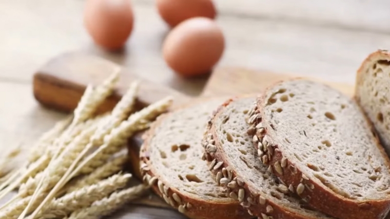 Slices of seeded whole grain bread on a wooden board with wheat stalks and eggs in the background