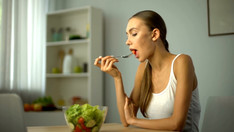 A woman in a white tank top eats a fresh salad at home, seated at a table with natural light in the background