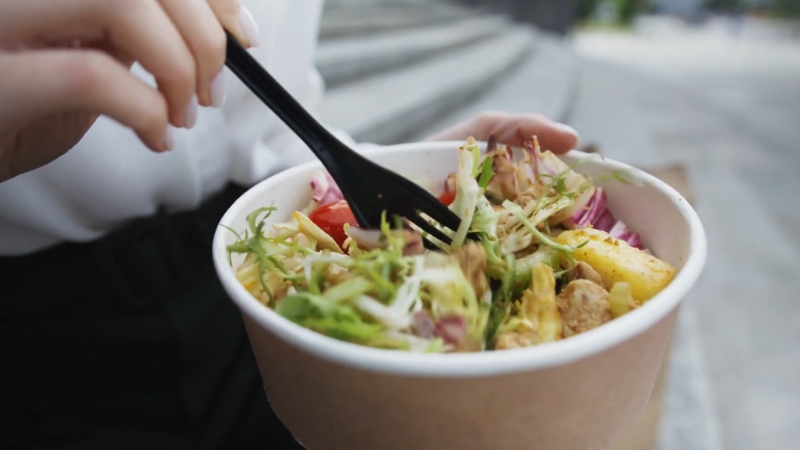 A person eats a fresh salad with greens, chicken, and vegetables from a paper bowl using a black plastic fork