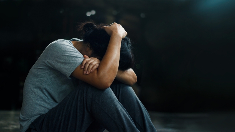 A person sits on the floor with their head down and arms wrapped around their knees