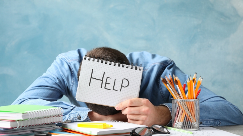 A person sits overwhelmed at a cluttered desk, holding a notepad that says “HELP,” showing a clear sign of burnout and the need for recovery